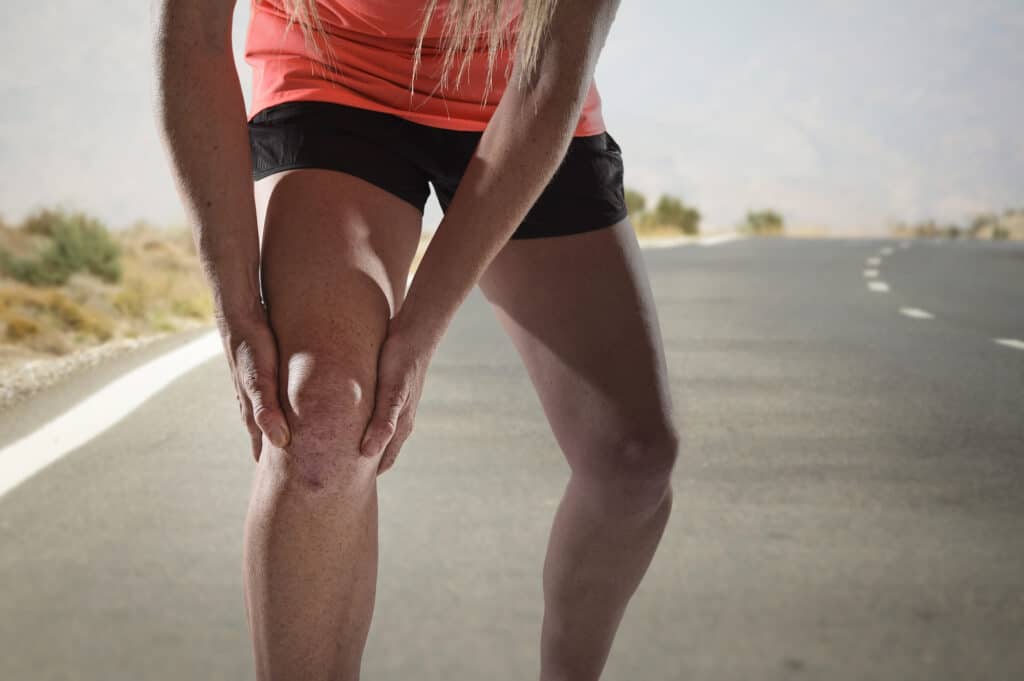 young sport woman with strong athletic legs holding knee with his hands in pain after suffering ligament injury during a running workout training on asphalt road background