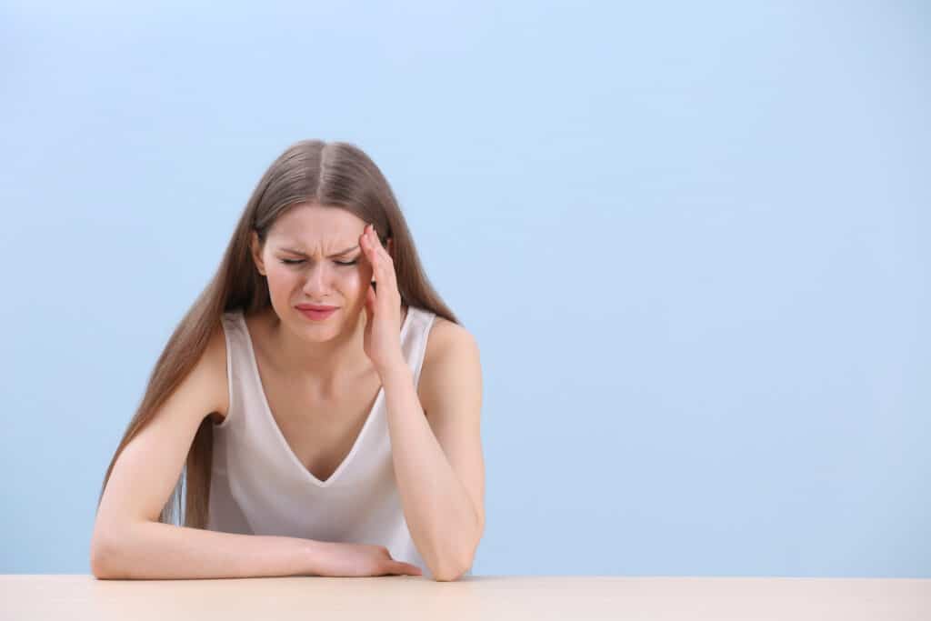Beautiful young woman suffering from headache while sitting at table, on color background